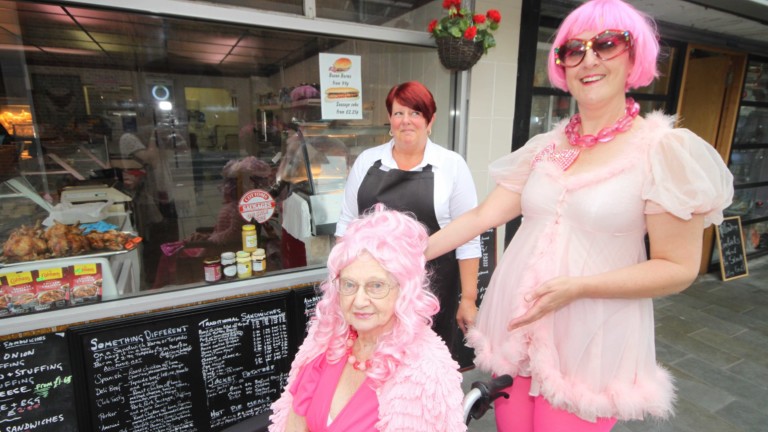Two people in pink wigs are in front of a takeaway window. One person sitting in a wheelchair wears a bright pink dress and soft pink cardigan. The other wears a pink fluffy dress, bright pink tights. and sunglasses. They gesture to point your attention to the person sitting, smiling widely. Behind the two people another stands smiling and looking at the two people in pink.