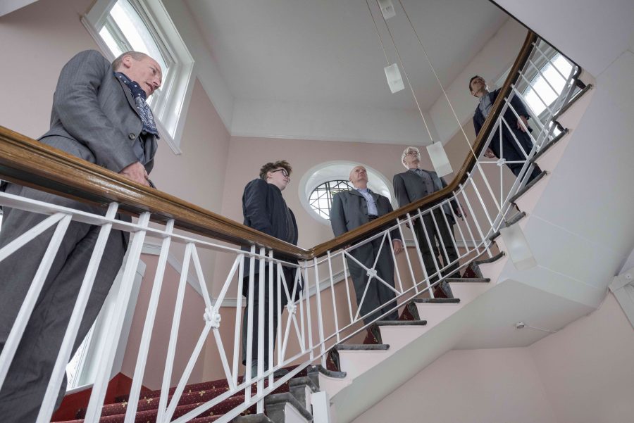 Five men in smart clothing stand at different heights on a winding staircase. They all face forward as the camera looks on from below.