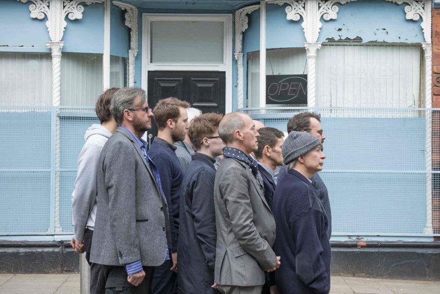 A group of men stand tightly grouped together in the middle of an empty road. They all face in one direction.