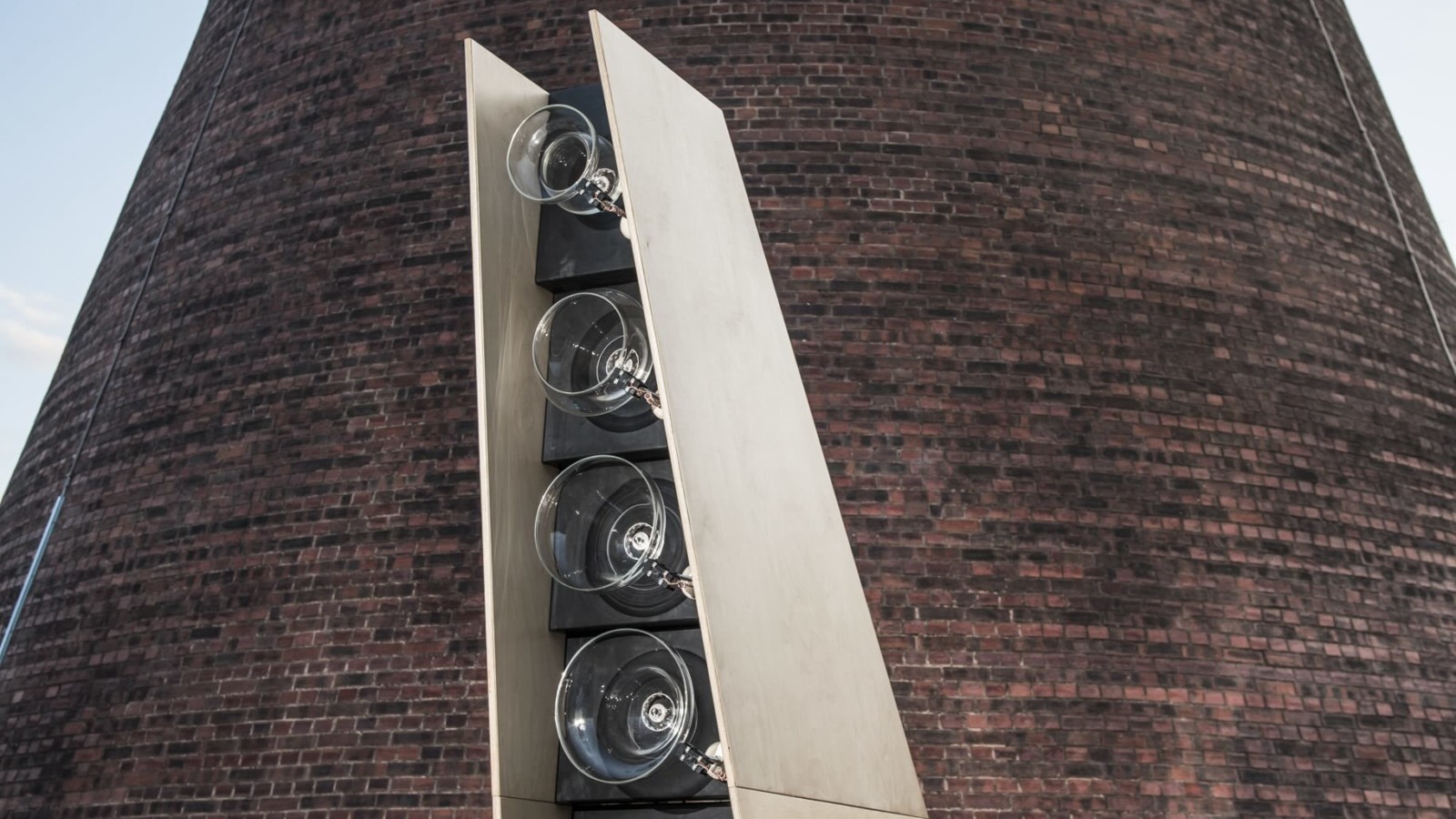 In front of a large conical building stands a structure around 2 meters tall. The structure is made out of sheets of naturally coloured wood and black wood. It is tapered and larger at the bottom than the top. On the front of the structure are 6 glass circles, in a vertical line. The glass circles get smaller the higher up the structure they reach.