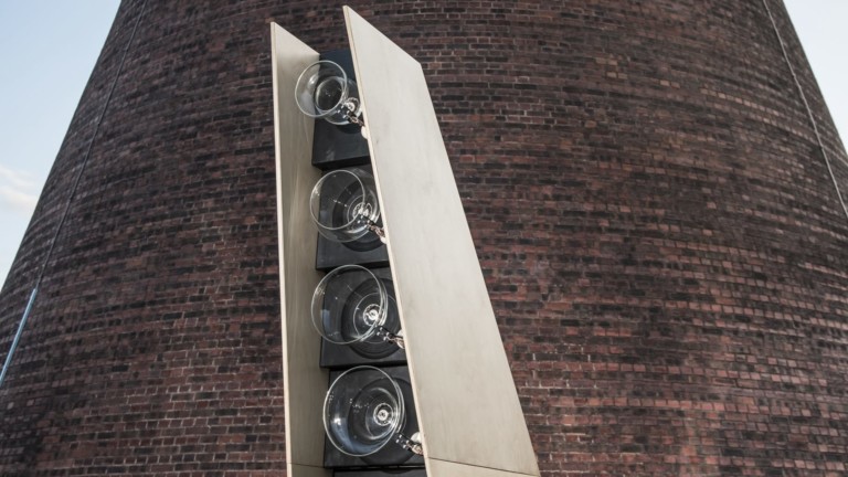 In front of a large conical building stands a structure around 2 meters tall. The structure is made out of sheets of naturally coloured wood and black wood. It is tapered and larger at the bottom than the top. On the front of the structure are 6 glass circles, in a vertical line. The glass circles get smaller the higher up the structure they reach.