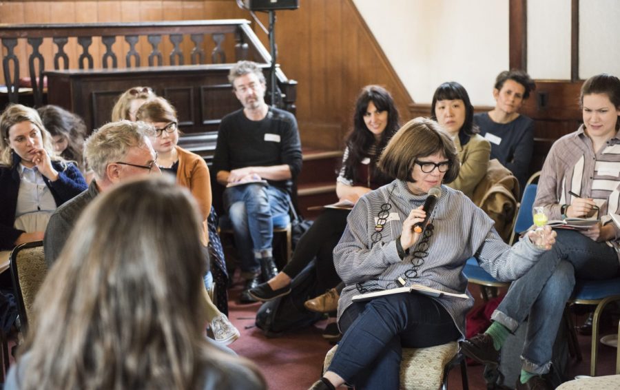 Artist Lois Weaver sits in the middle of an audience of poeple holding a microphone and gesturing with her hands.