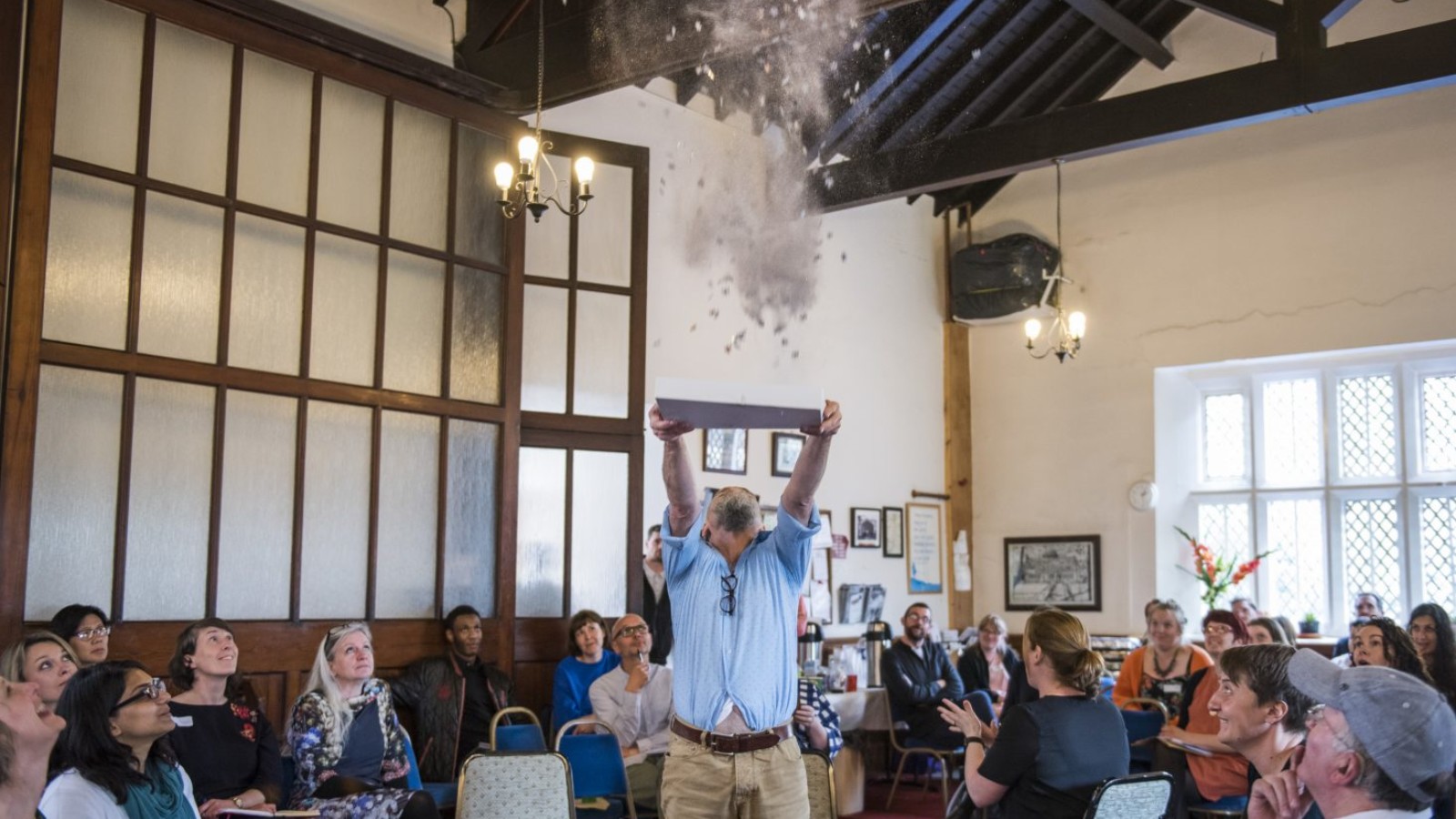 In the middle of a large room with wooden beams and dividers a man stands throwing a cloud of jigsaw pieces and glitter in to the air above his head. An audience look in to the air towards the cloud, some smiling.