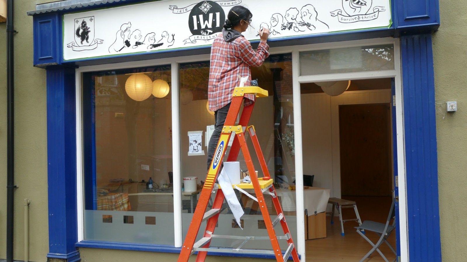 A person with dark hair in a ponytail stands on a ladder in front of a shop doorway and is painting a black and white sign. In the middle of the sign there is a crest which reads 'iwi'.