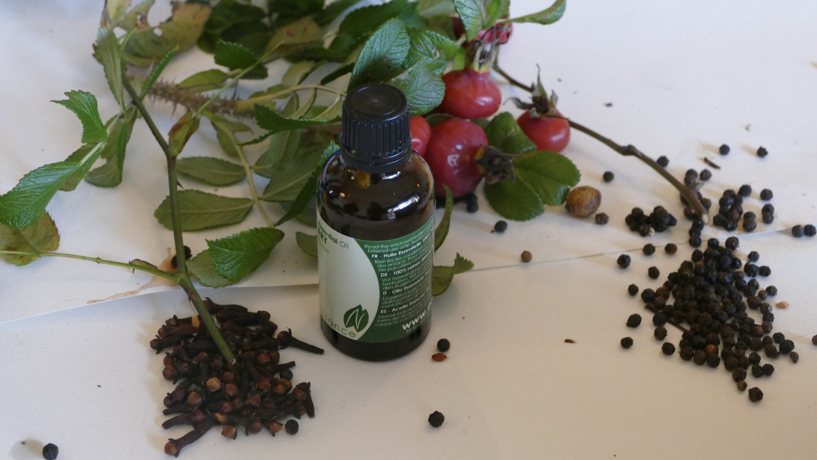 On a white table a collection of herbs, spices and botanicals are arranged around an essential oils bottle.