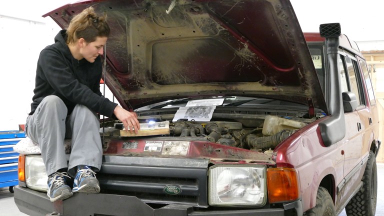 Artist Dina Rončević sits on the front of a maroon coloured Land Rover with the bonnet open.