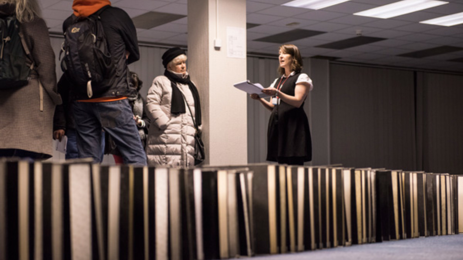 Artist Michelle Browne stands in a black dress in a room lit by fluorescent lights. She is holding a piece of paper in her hands and speaking to an audience of people.