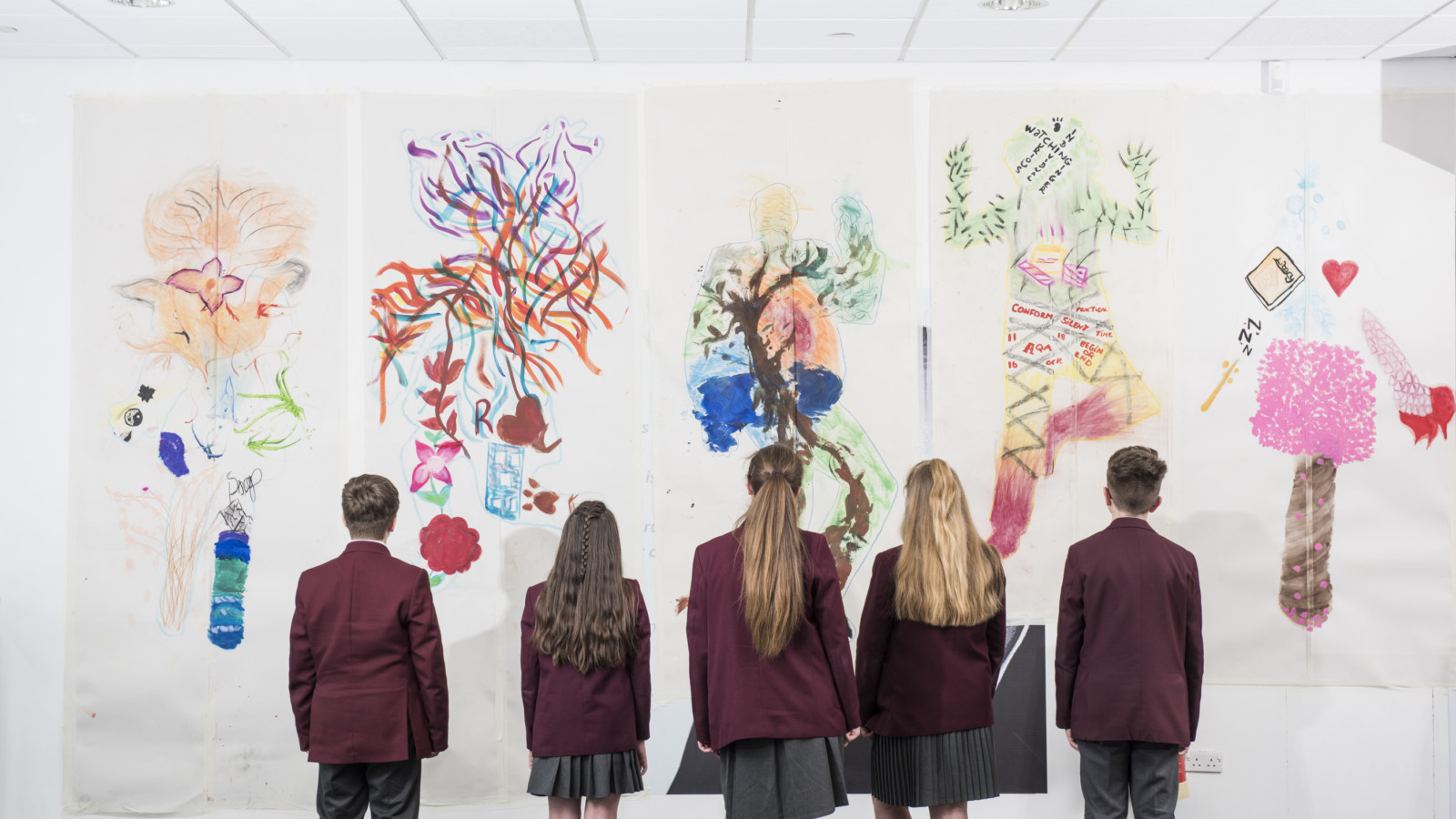A group of young people stand facing a collection of brightly coloured artworks. The young people are all wearing maroon blazes, some of have long hair and some have short, cropped hair.