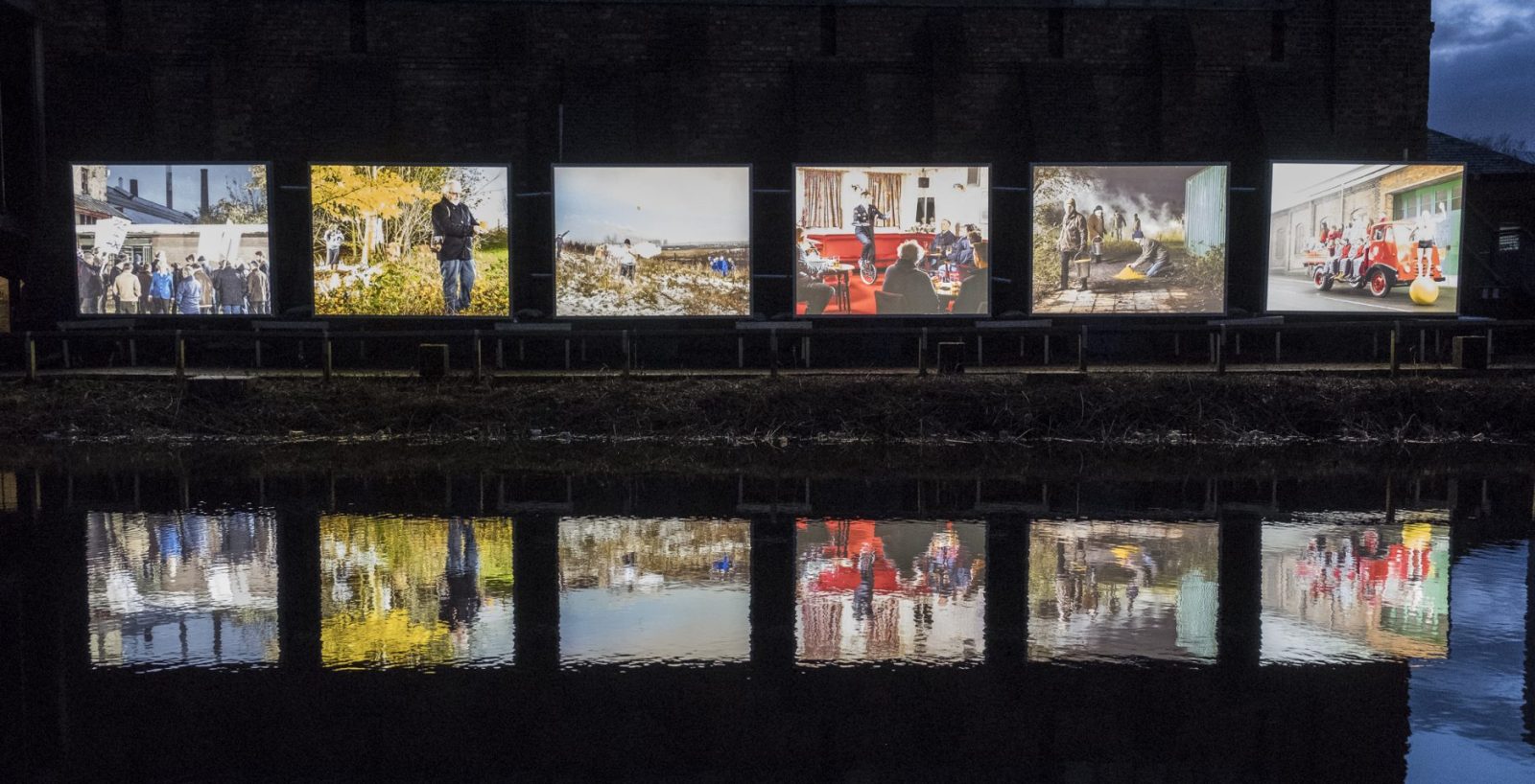 6 large light boxes with a different image in each stand in a row next to a canal. The canal water reflects the images. It's dark outside.