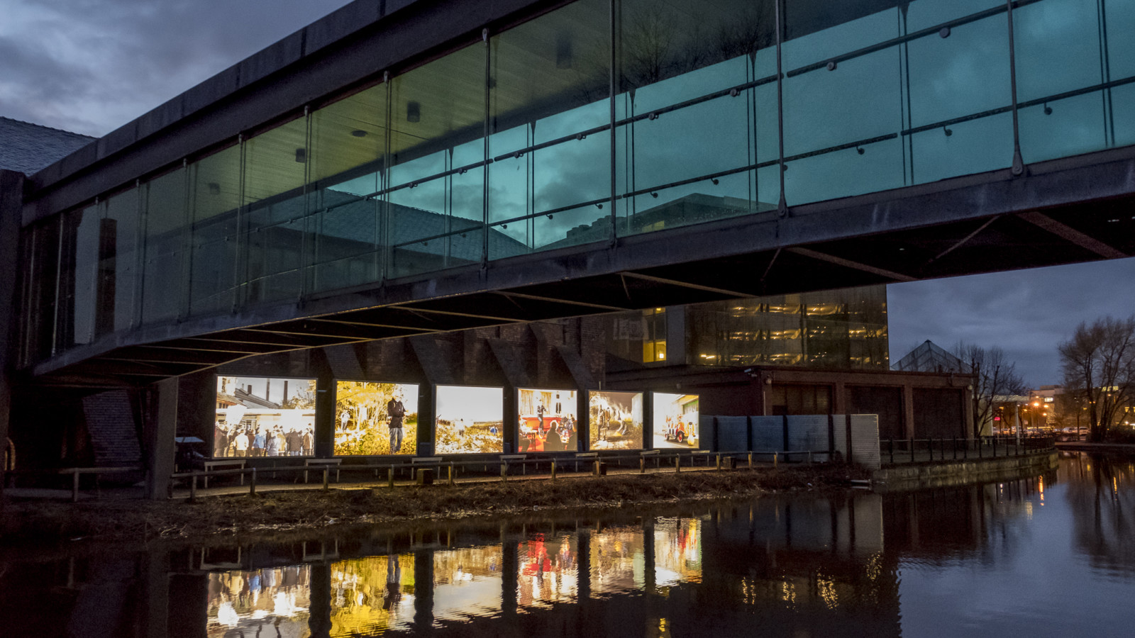 Six large light boxes with a different image in each stand in a row next to a canal. The canal water reflects the images. It's dark outside. A glass covered bridge is suspended over the water.