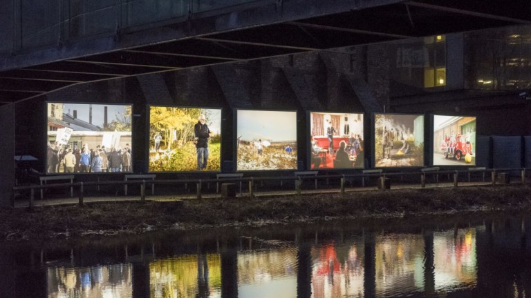6 large light boxes with a different image in each stand in a row next to a canal. The canal water reflects the images. It's dark outside.
