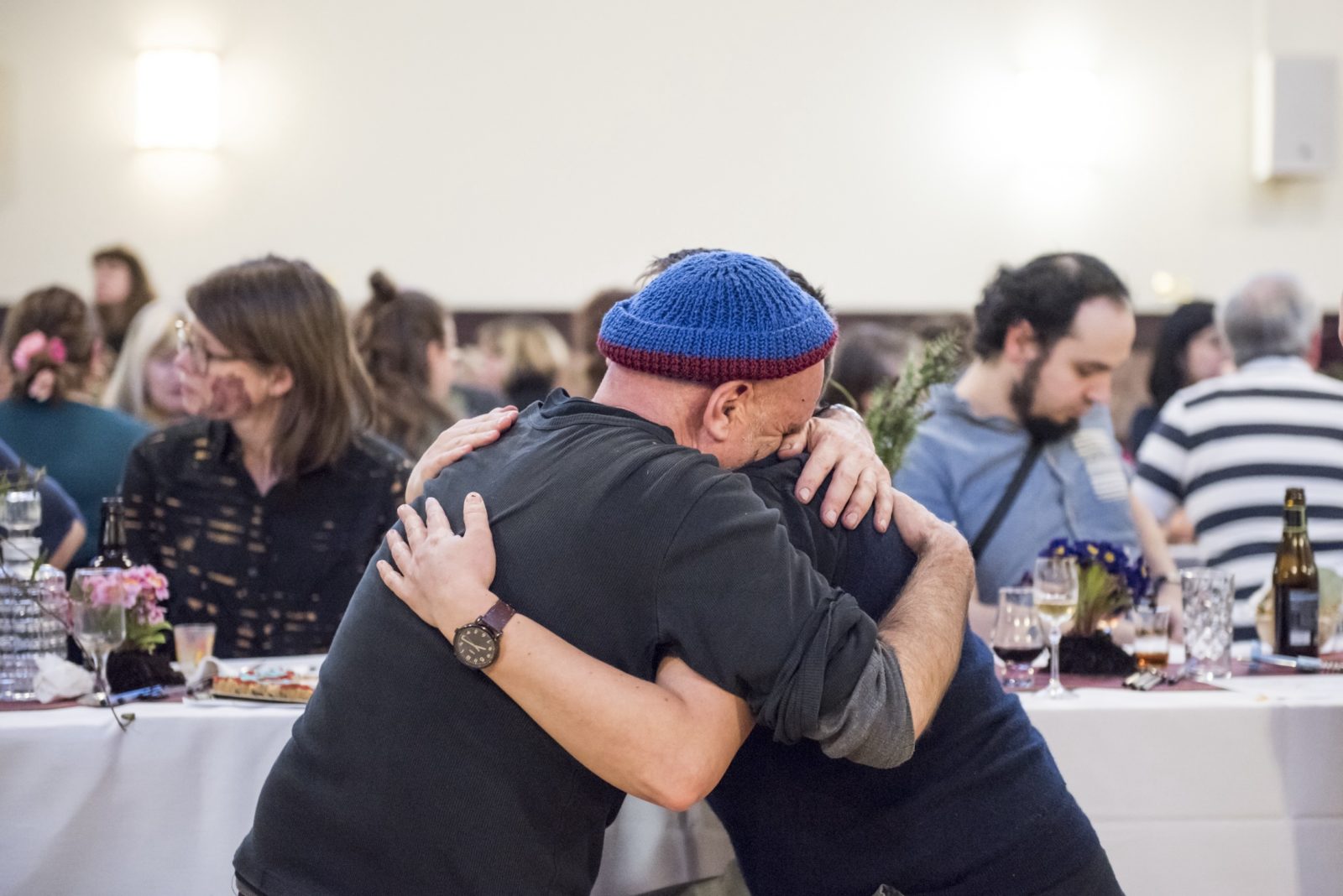 Two people sitting at a table hug intensely. One wears a blue hat and buries their head in the shoulder of the other. Behind them the room is full of people sitting at long tables.