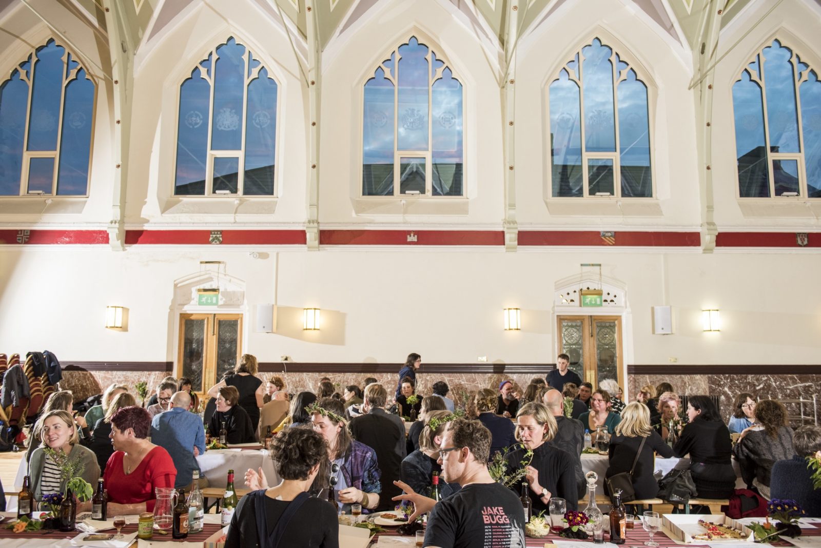 A large group of people gather around two long tables in the centre of a large hall. The walls of the hall are white and there are 5 large and ornate windows that reveal a dusky sky.