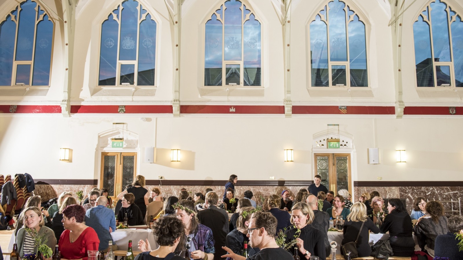 A large group of people gather around two long tables in the centre of a large hall. The walls of the hall are white and there are 5 large and ornate windows that reveal a dusky sky.