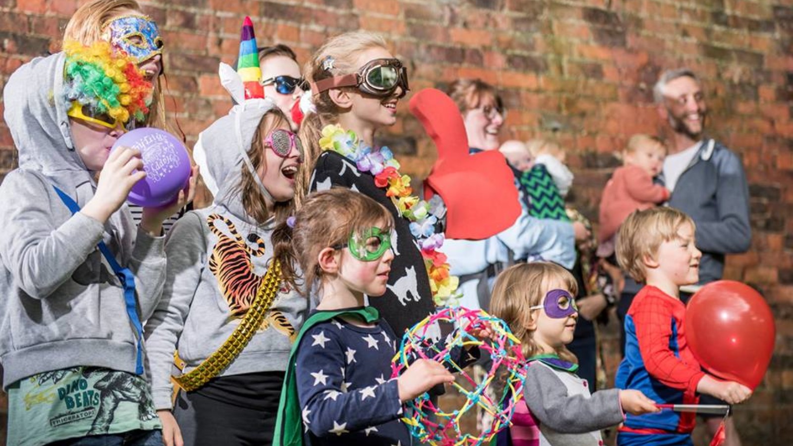 A group of children and adults stand in front of a brick wall wearing superhero masks and capes.