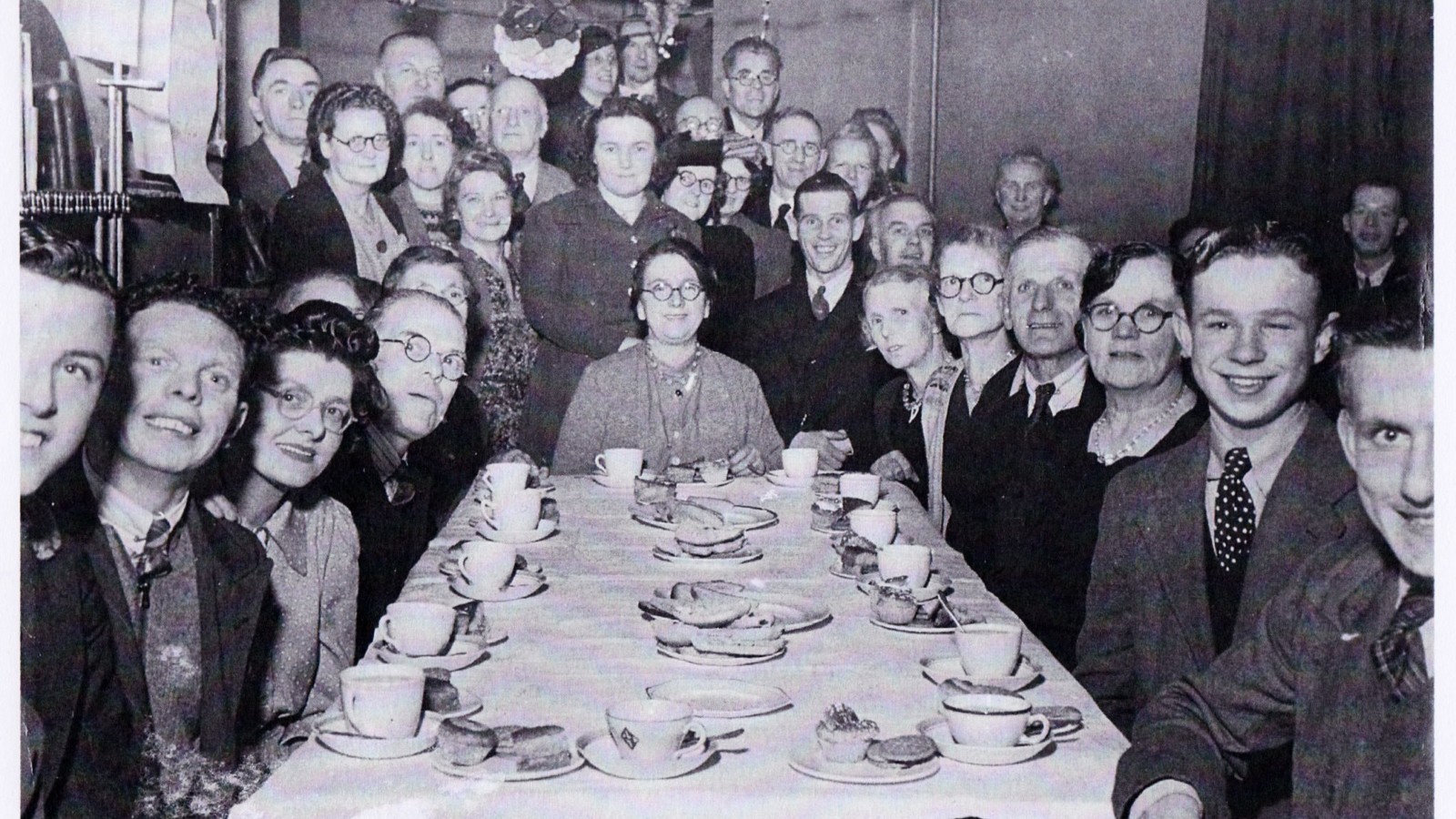 A black and white photograph of a large number of people sitting and standing around a table, posing for a photograph. The table is set with teacups and saucers.