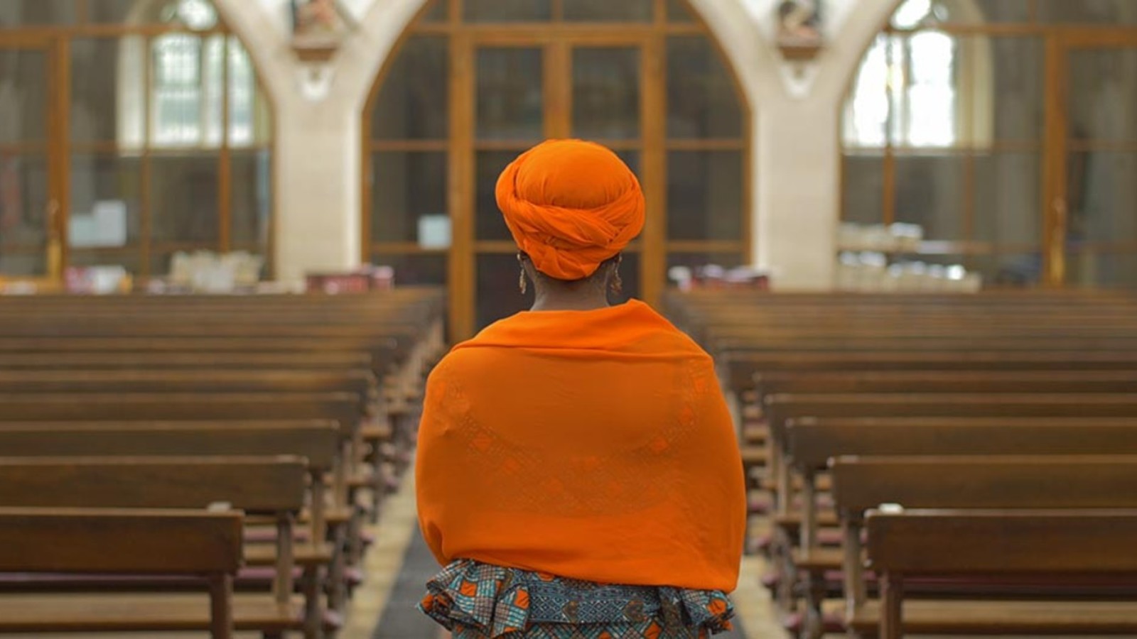 The back of a woman stands in the middle isle of an empty church wearing a bright, orange headscarf and scarf around their shoulder.