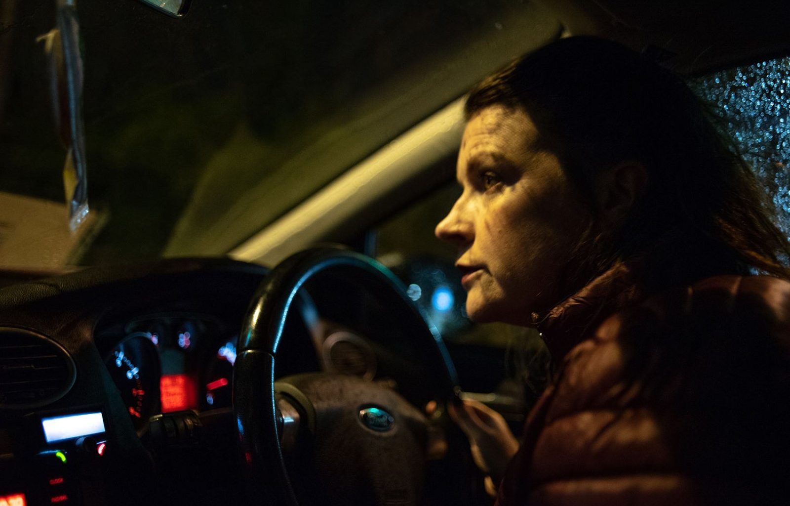 A woman sits at the wheel of a car looking out of the windscreen. The car dash lights are on and it's dark outside, a streetlight lights the woman's face.