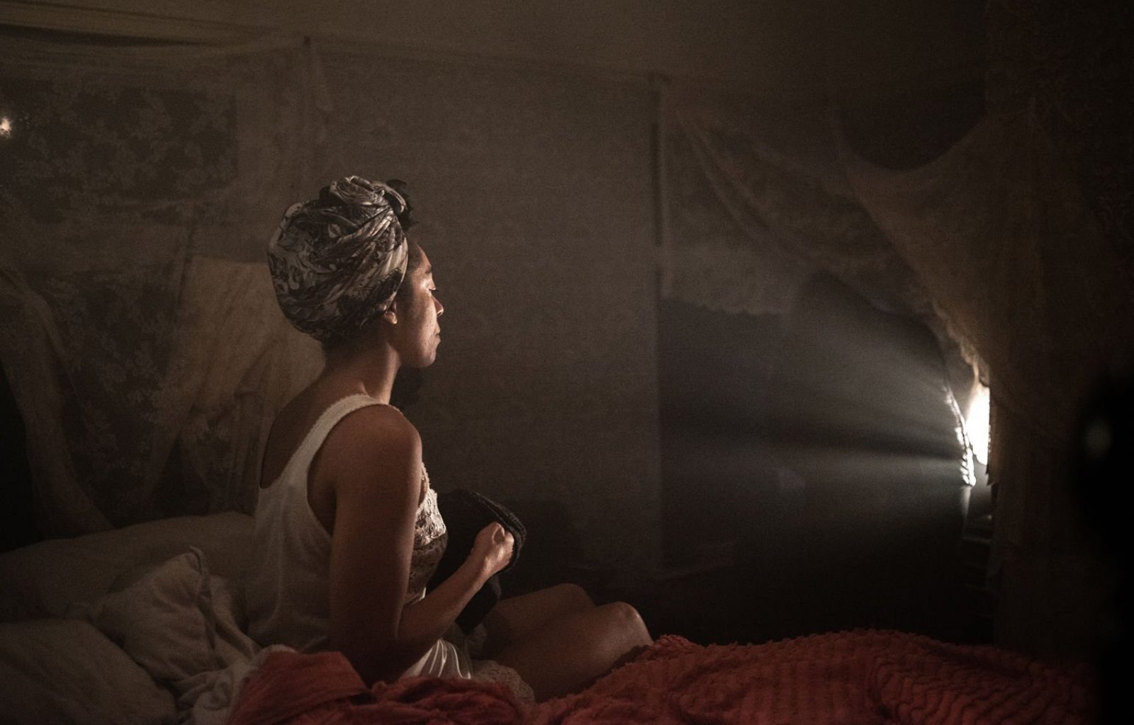 A woman sits on an unmade bed, in a nightgown and her hair tied up in a scarf. We see her from the side. She sits in a dark room and is lit from the front by a bright lamp. She is looking at the light.