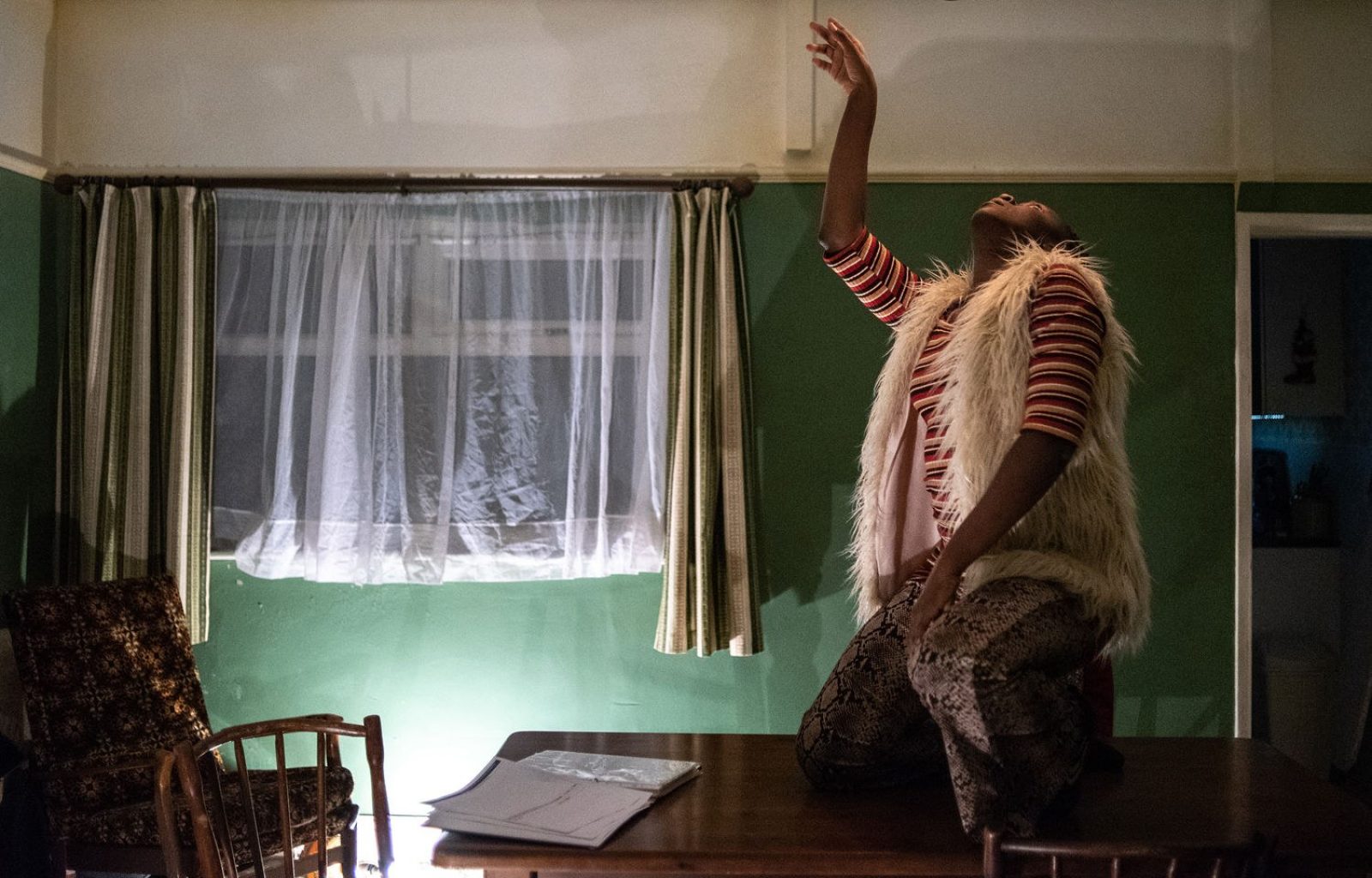 In a green and white room a woman dances on a dark wooden table. The woman is dressed in snake print trousers and a white fluffy vest  and kneels on top of the table with her hand in the air.