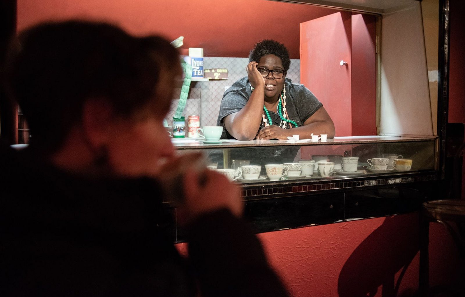 A woman leans on a glass counter in a red room. Inside the counter is a collection of teacups. Out of focus, in the foreground, a person with short hair sips from a teacup.