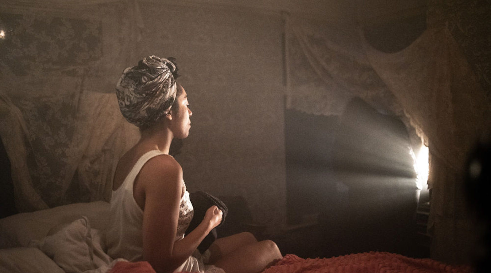 A woman wearing a nightdress and a hair wrap is sitting on the edge of a dishevelled bed. She is looking off camera towards a patch of light in the corner of the room. The room is dark and the light shines on her face.