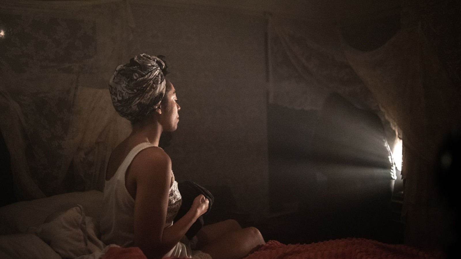 A woman sits on an unmade bed, in a nightgown and her hair tied up in a scarf. We see her from the side. She sits in a dark room and is lit from the front by a bright lamp. She is looking at the light.