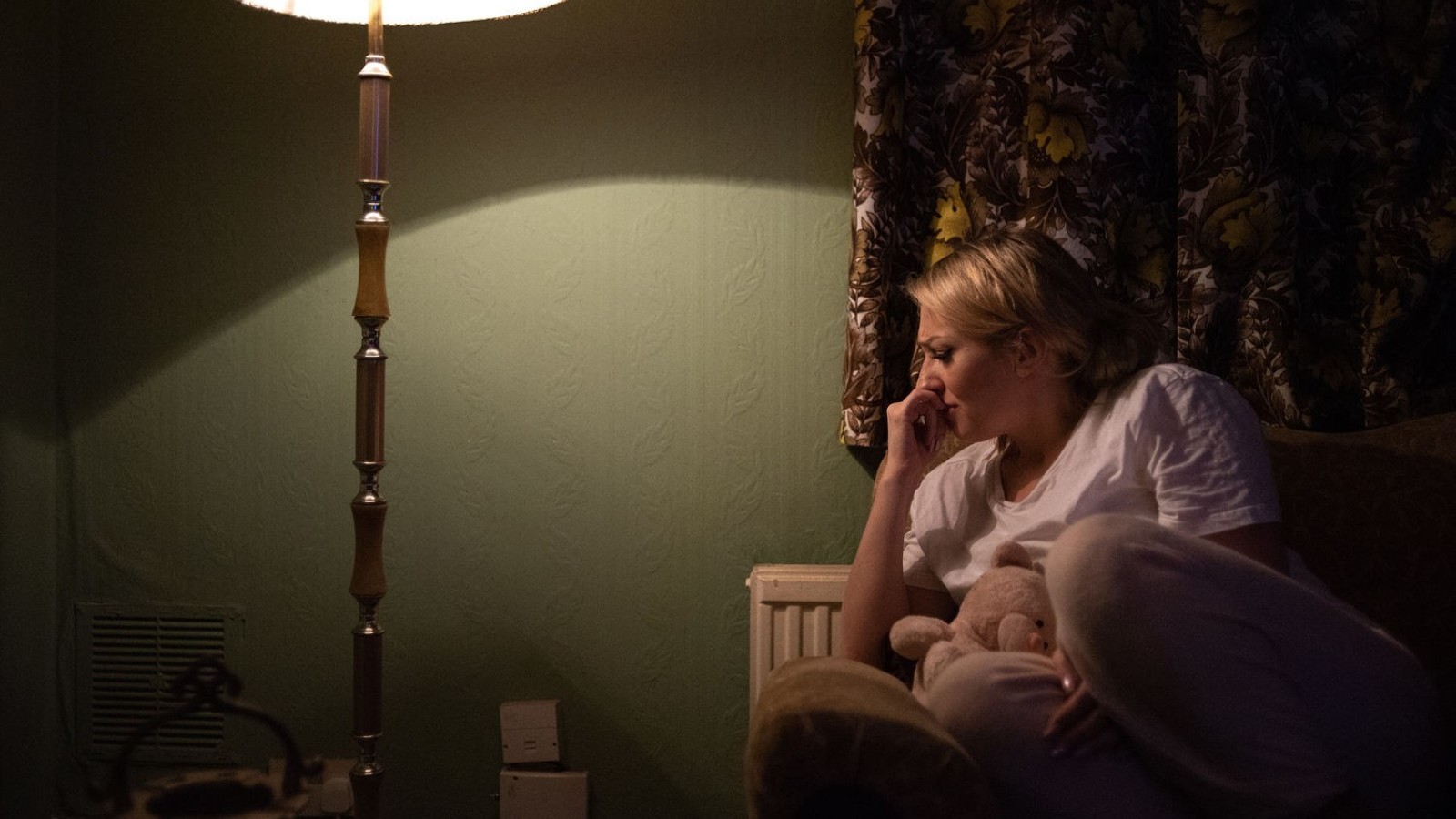 A blonde haired woman sits in a dark living room, lit by a tall standing lamp. Wearing pink pyjamas, she is curled up on the couch. She holds a teddy close with her left arm and her right hand is curled to her face. She looks sad, in distress.