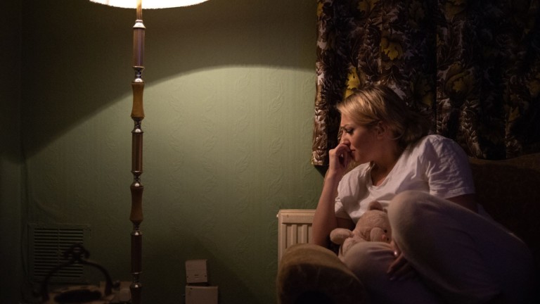 A blonde haired woman sits in a dark living room, lit by a tall standing lamp. Wearing pink pyjamas, she is curled up on the couch. She holds a teddy close with her left arm and her right hand is curled to her face. She looks sad, in distress.