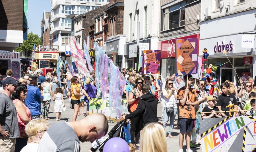 It is a summers day on a busy pedestrianised high street. Crowds of people are watching as a parade of people walk by holding up colourful banners and flags.