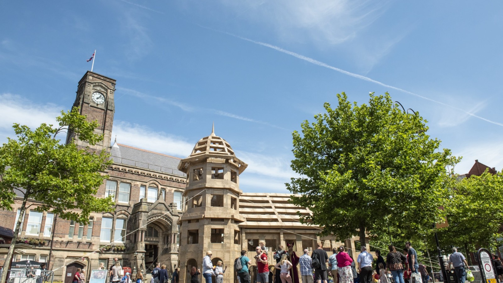 A crowd of people are gathered underneath a huge castle made of cardboard. It is a sunny warm day with blue skies. The castle sits in front of a large Town Hall building.