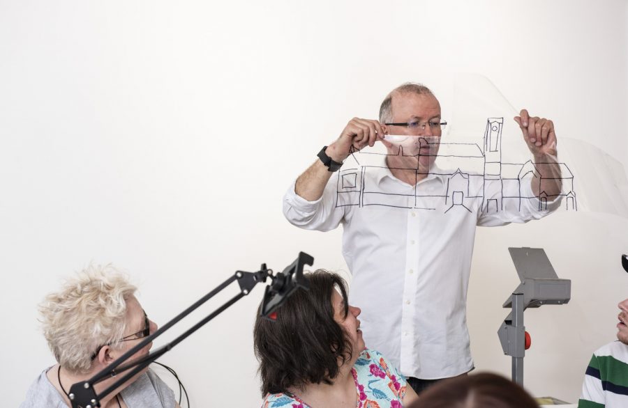 A person with short, light hair, wearing a white shirt stands holding a piece of clear plastic with a drawing of a building in black marker up in the air. You can see an overhead projector in the bottom corner.