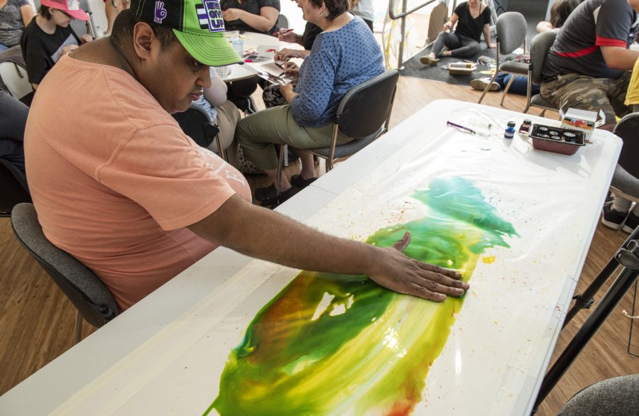 In a busy room a person wearing a cap and t-shirt sits at a table. On the table is multiple colours of paint enclosed between clear plastic. The person is rubbing the paint around underneath the clear plastic, mixing the colours together.