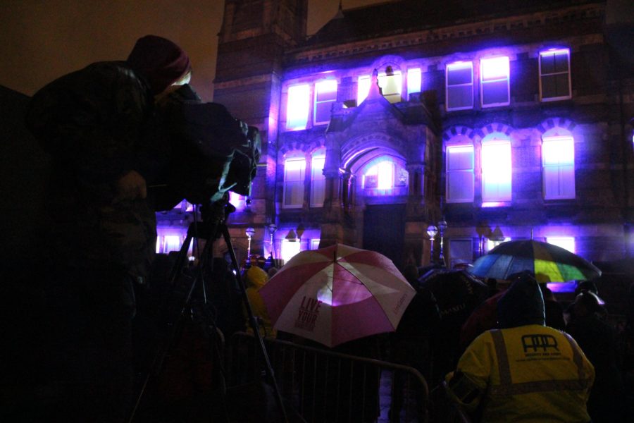 St Helens Town Hall has it's windows lit up with bright light purple projections against a night sky. An audience stands in front of the building with umbrellas.