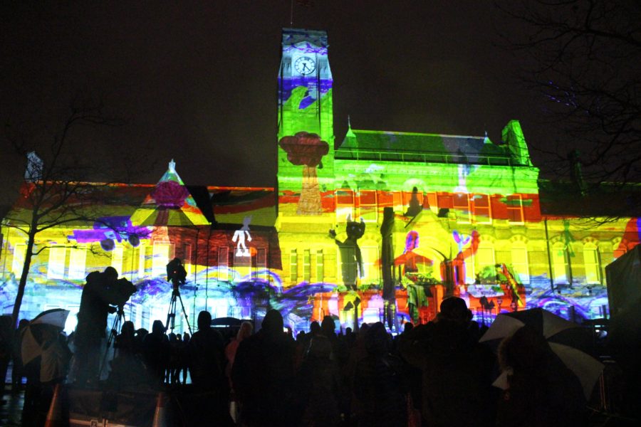 St Helens Town Hall is lit up with a huge multicoloured projection against a night sky. In front of the building we can see a large audience in silhouette.