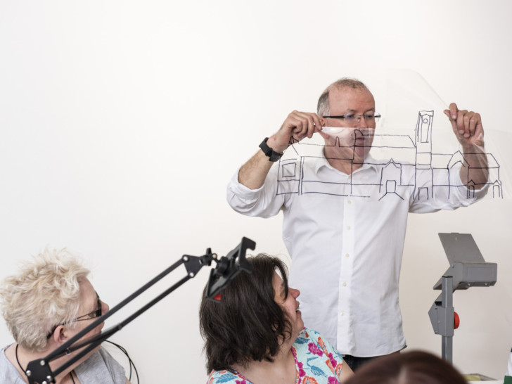 A person with short, light hair, wearing a white shirt stands holding a piece of clear plastic with a drawing of a building in black marker up in the air. You can see an overhead projector in the bottom corner.