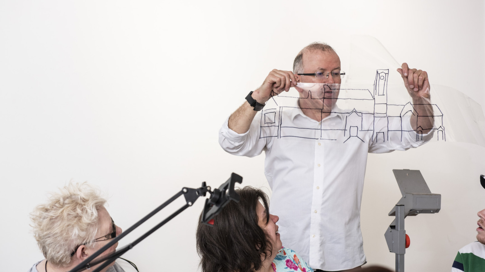 A person with short, light hair, wearing a white shirt stands holding a piece of clear plastic with a drawing of a building in black marker up in the air. You can see an overhead projector in the bottom corner.