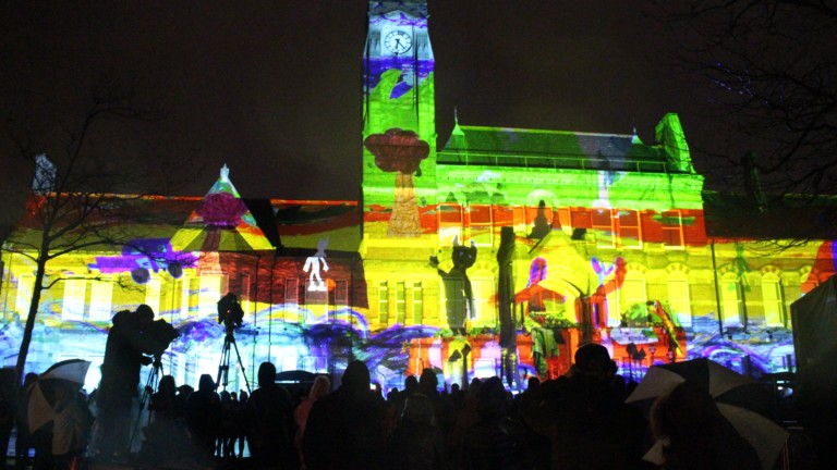 St Helens Town Hall is lit up with a huge multicoloured projection against a night sky. In front of the building we can see a large audience in silhouette.