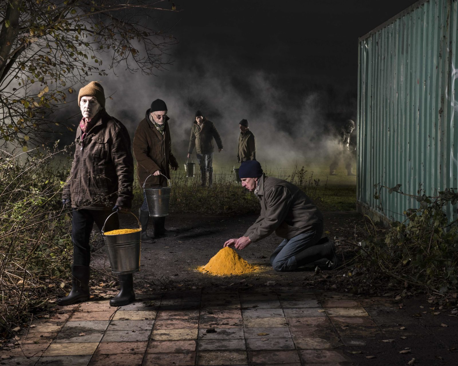 Five men dressed in winter coats and hats stand and kneel on patio slabs and a field. One tends to a pile of mustard coloured powder, the others carry metal buckets full of mustard coloured tablets, there is a gorilla and smoke in the background.
