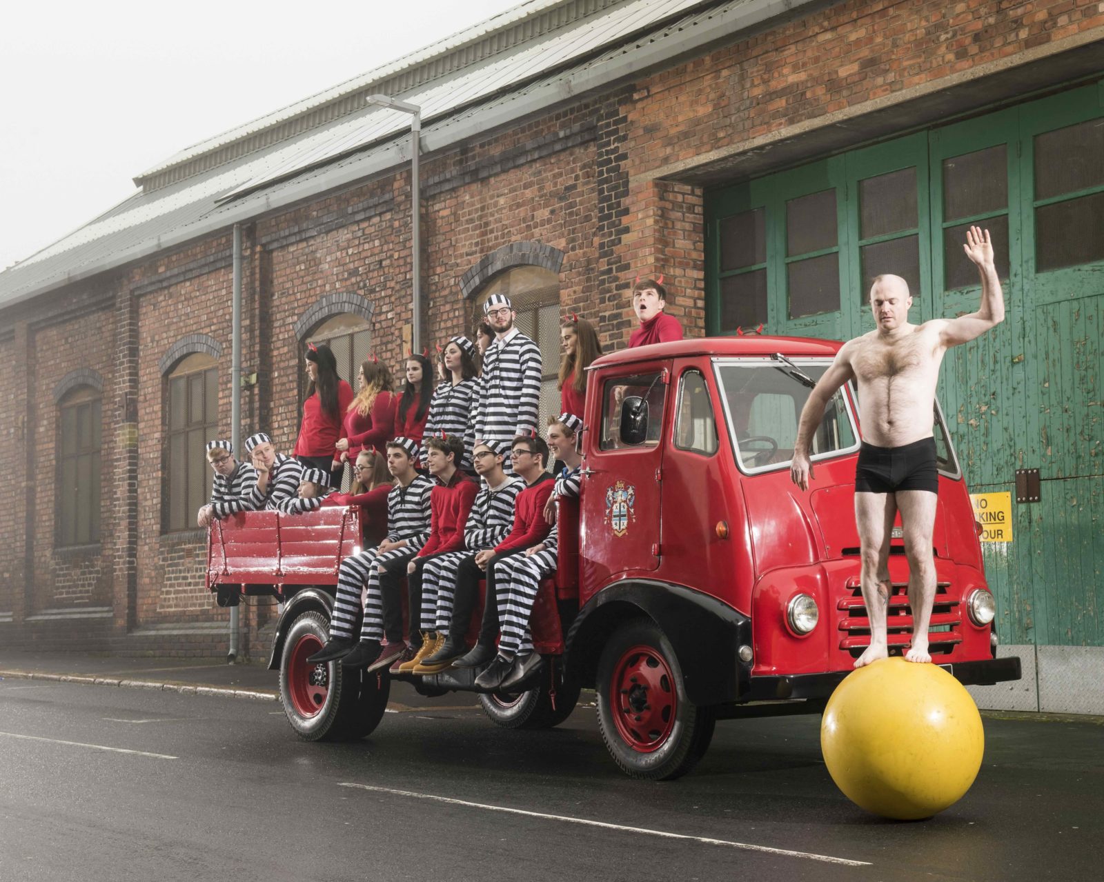 In front of a large green garage door and red brick wall a bright red, classic flat bed truck stands in the road. On the back of the truck sit and stand a group of people. Some wear striped outfits and some wear red outfits with fake horns. At the front of the truck and man balances on a large yellow ball wearing only black shorts.