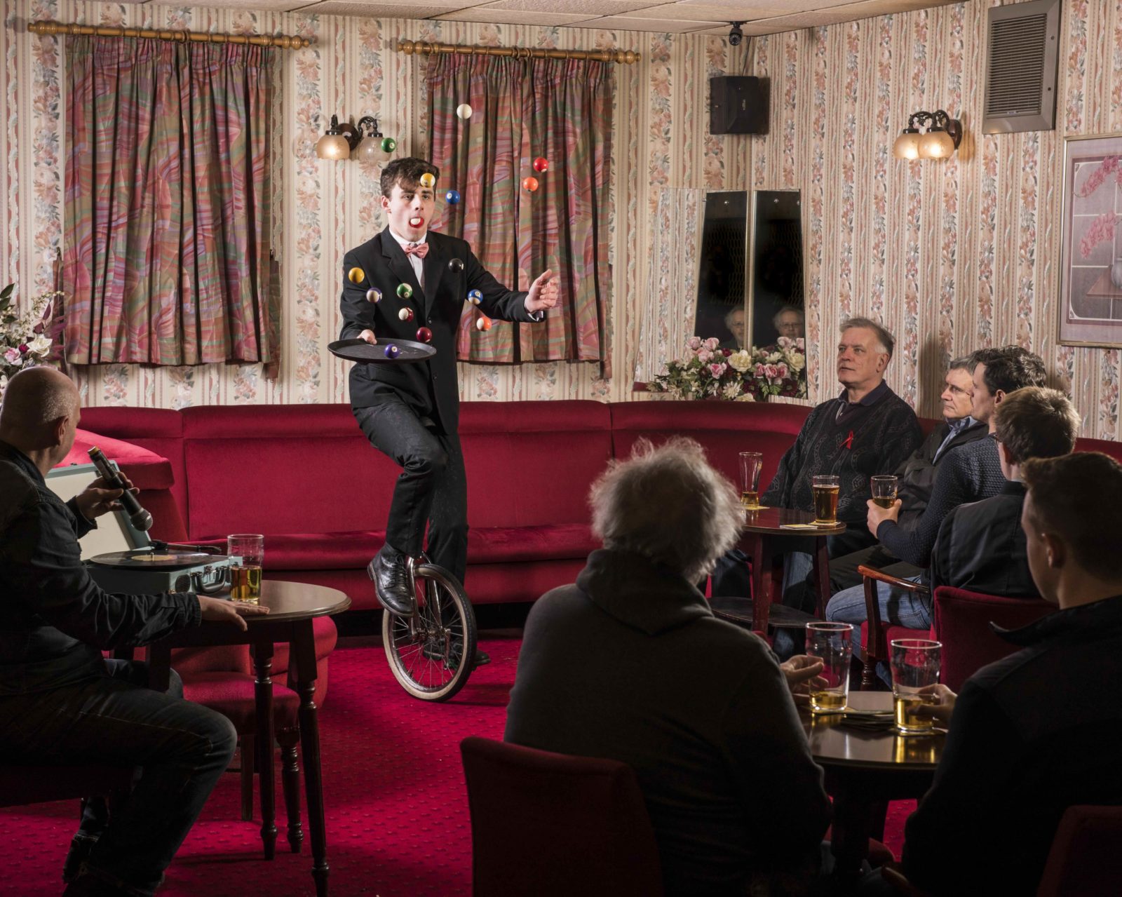 A group of men sit in a pub lounge. The seats and carpet are vibrant, deep red. There are pink and red flowery curtains and walls. All the men look towards the centre of the room, where a young man rides a unicycle, holding a bar tray and juggling a set of snooker balls.