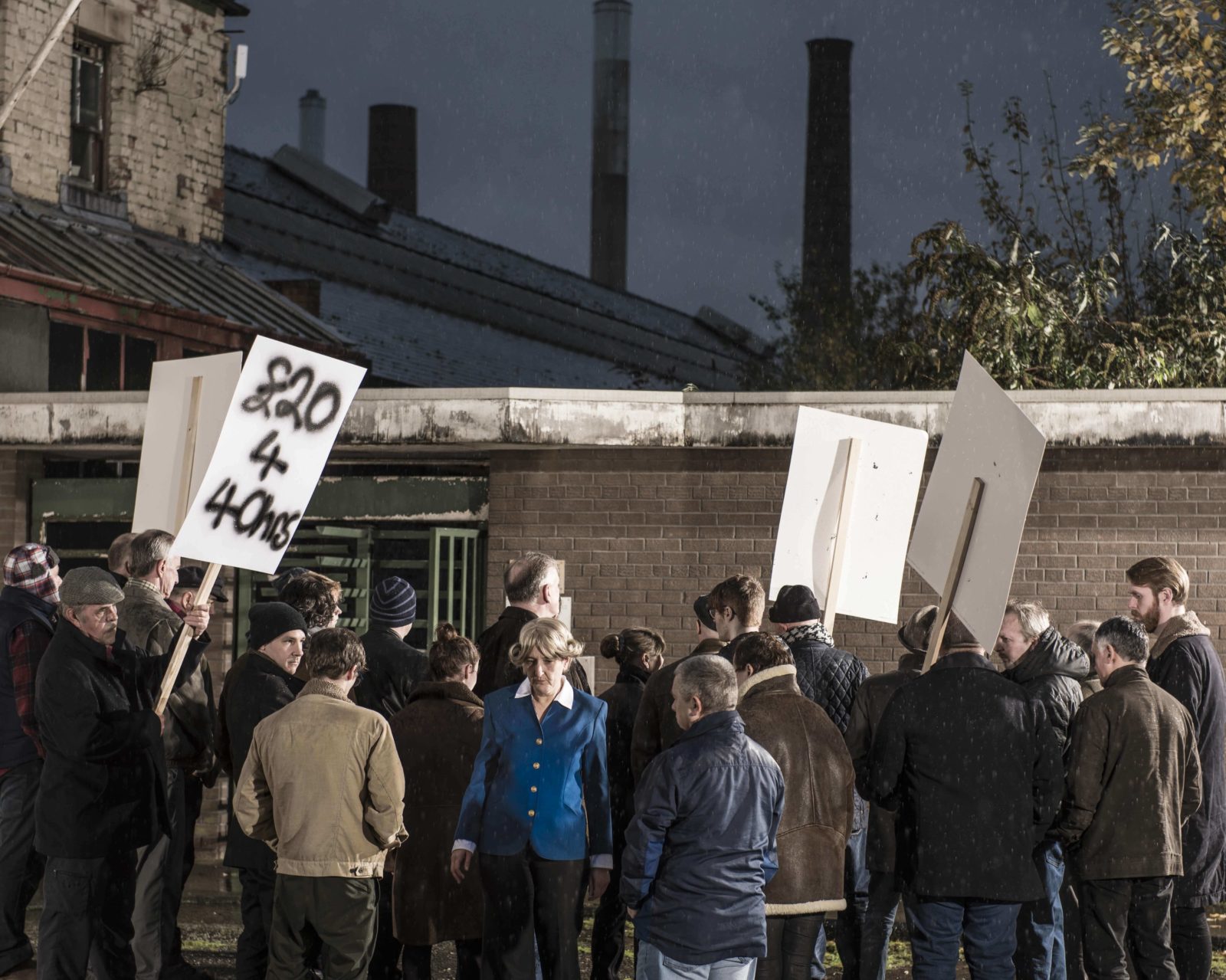 In front of a factory turnstile a crowd of people gather facing away from us. Some are holding placards. One placard reads '£20 for 40 Hours". In the middle of the crowd a woman in a blue jacket walks towards us, looking nervous.
