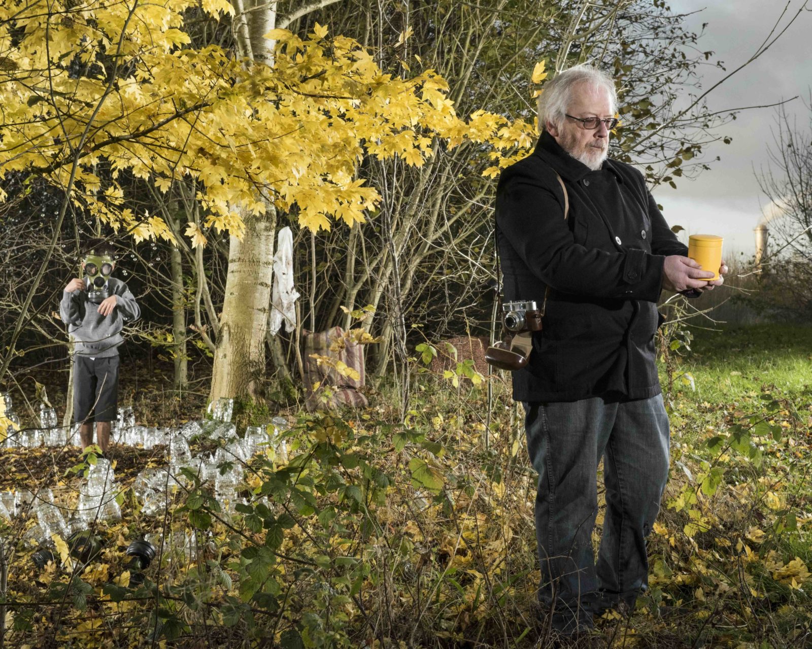 In a field next to a line of trees an man with light hair stands holding a ceramic yellow pot, with a camera hung over his shoulder. On the skyline, in the distance is a factory chimney with smoke coming out. Next to the man is a tree with bright yellow leaves. Underneath the tree a child in school uniform wears a gas mask and is surrounded by old fashioned pint glasses.