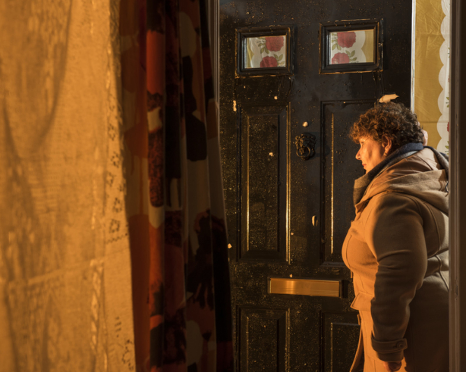 Bathed in yellow light, a woman wearing a brown coat stands at an open front door gazing out. The yellow light comes from outside, the house is otherwise in darkness.