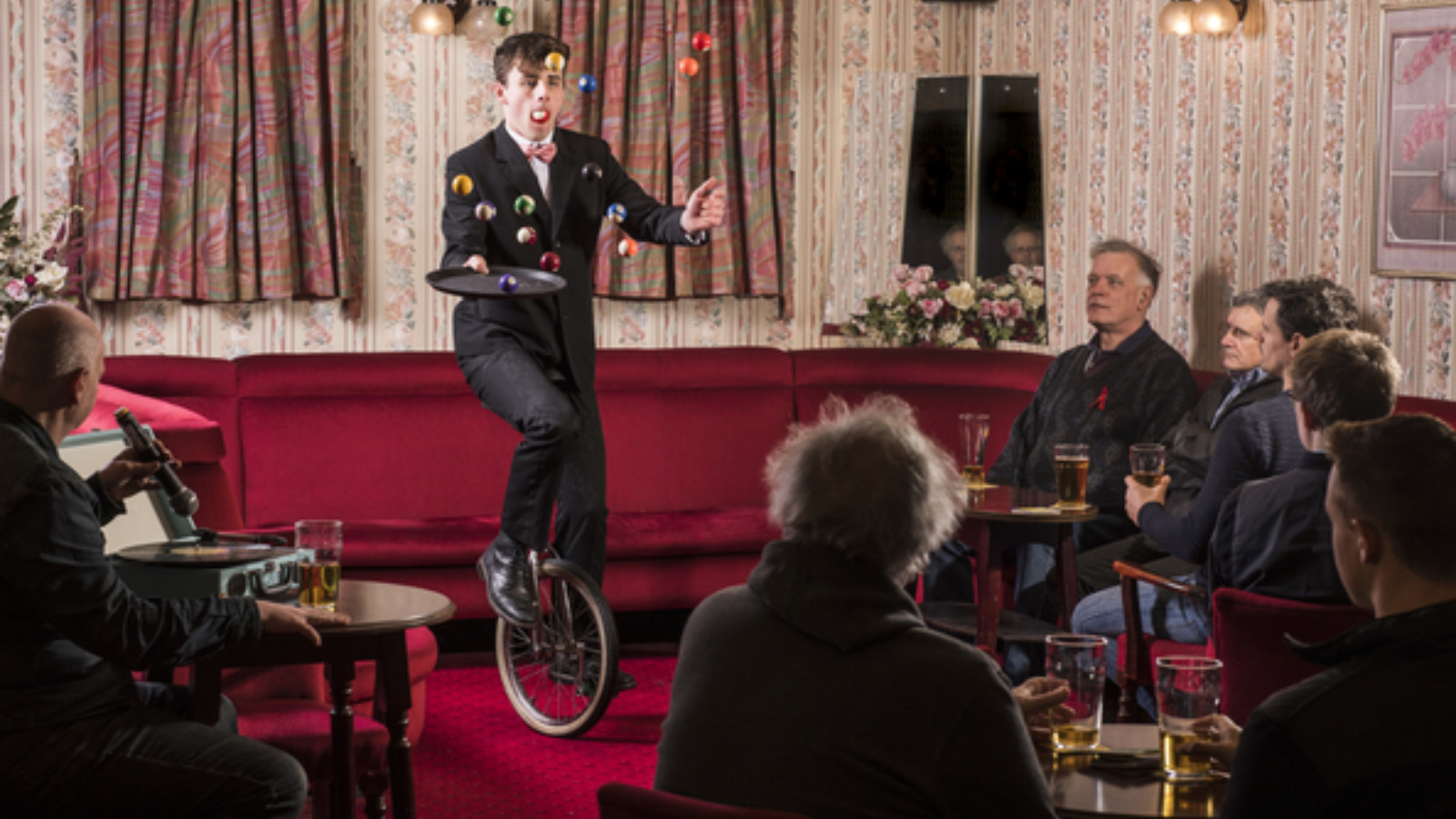 A group of men sit in a pub lounge. The seats and carpet are vibrant, deep red. There are pink and red flowery curtains and walls. All the men look towards the centre of the room, where a young man rides a unicycle, holding a bar tray and juggling a set of snooker balls.