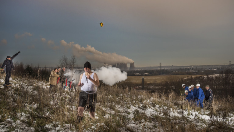 A group of men stand on a hillside, one is wearing a white vest, dark shorts and black beanie and is surrounded by a puff of smoke. Another throws a wreath towards a group of men in blue overalls and white hard hats.