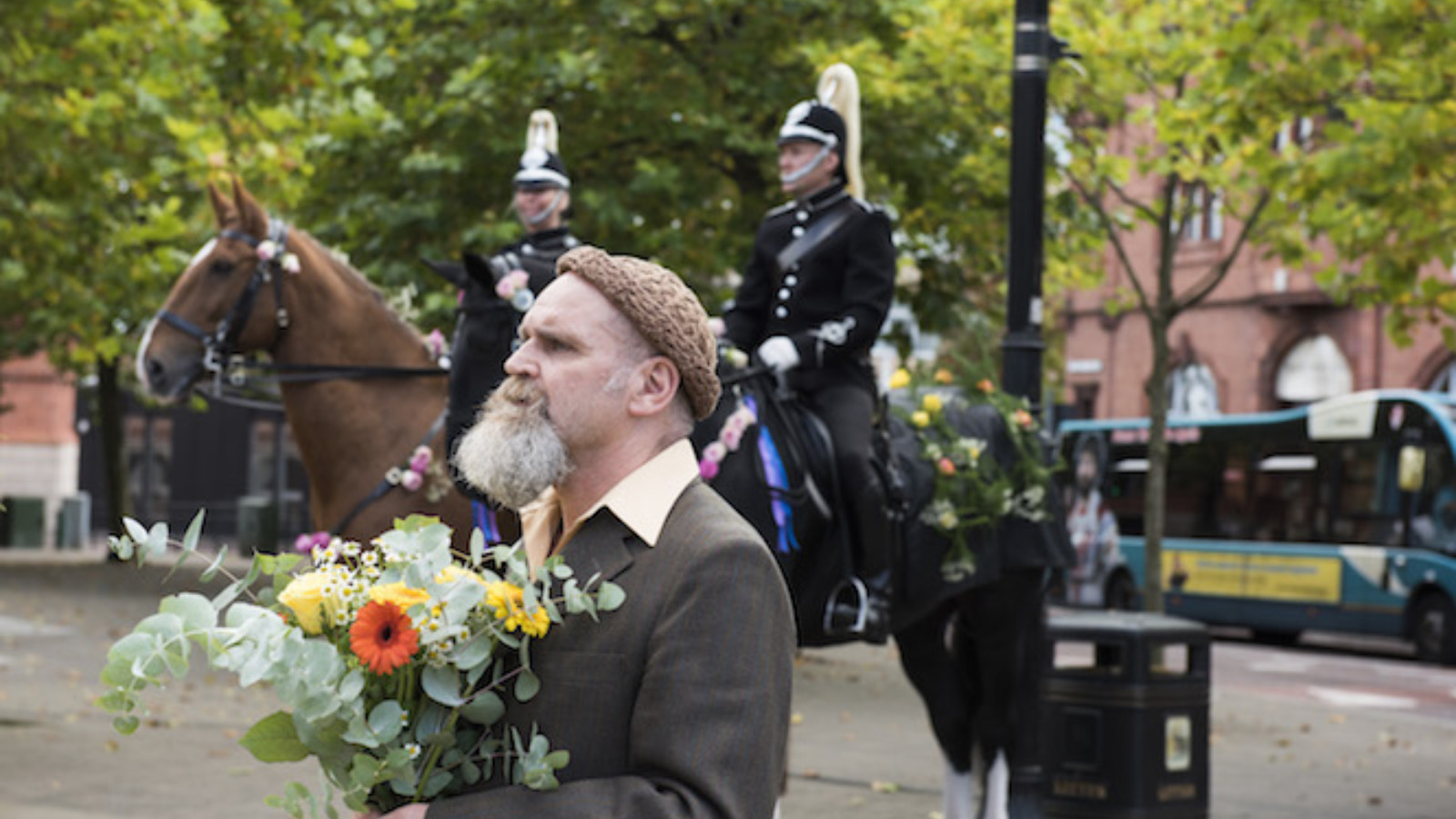 Artist Mark Storor stands in a community square wearing a brown suit and holding a large bunch of flowers. Behind Mark two mounted police officers sit on horses adorned with roses and greenery.