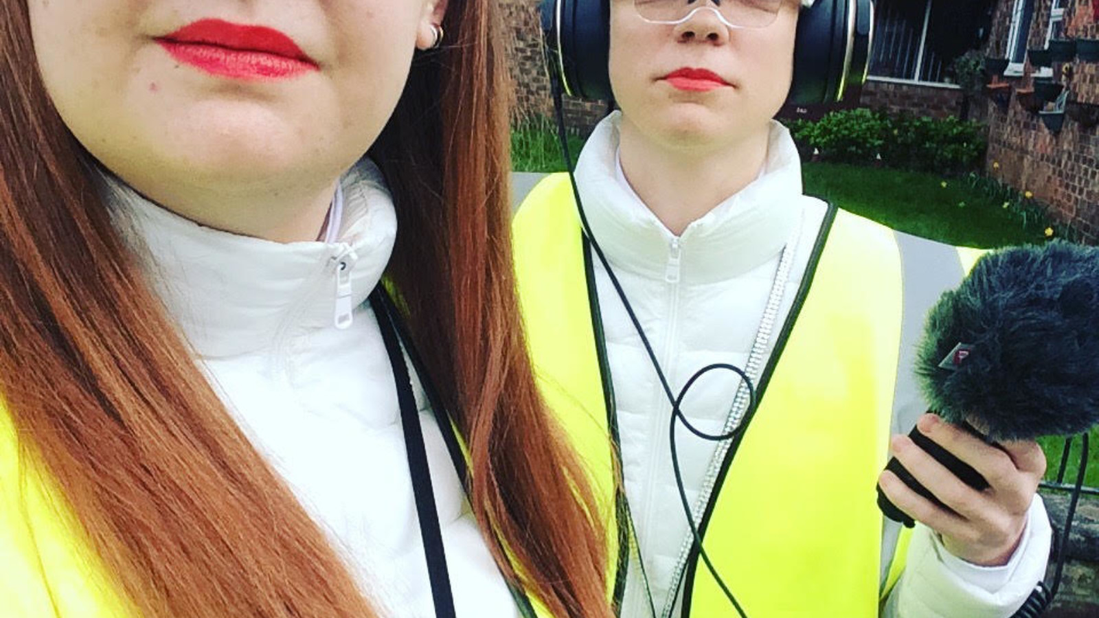 Rosana Cade and Sian Baxter stand in high visibility vests, wearing goggles and headphones, carrying a microphone.
