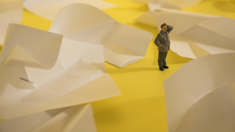 A miniature model of a person holding their hand on their head stands amongst white paper which is folded in to different shapes.