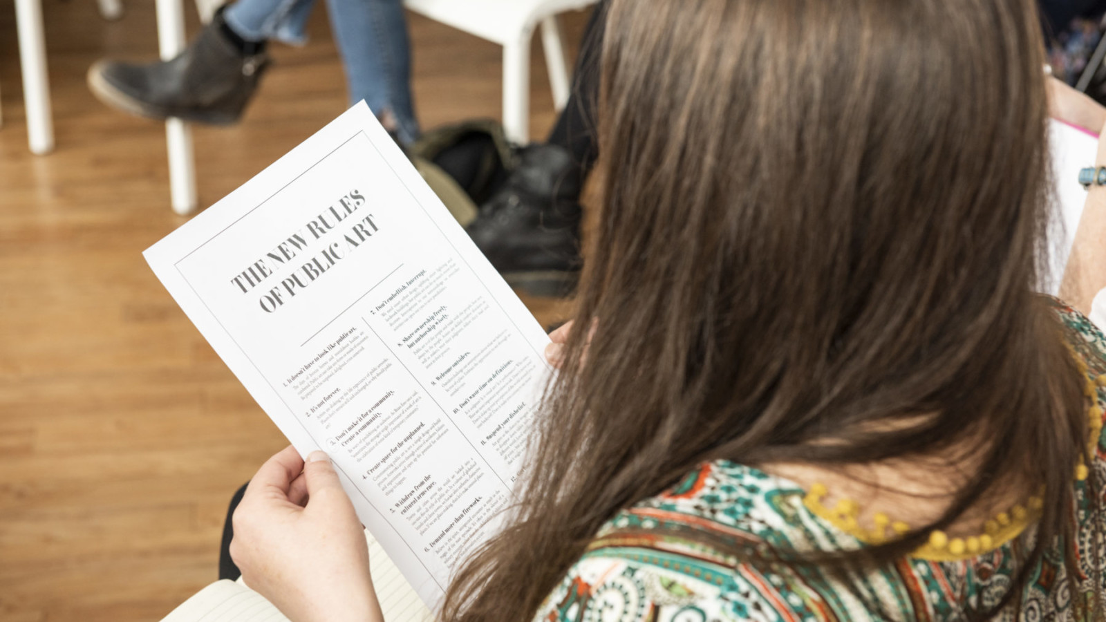 We look over the shoulder of a woman with long dark hair. She is reading a piece of paper with the title 'The New Rules of Public Art'